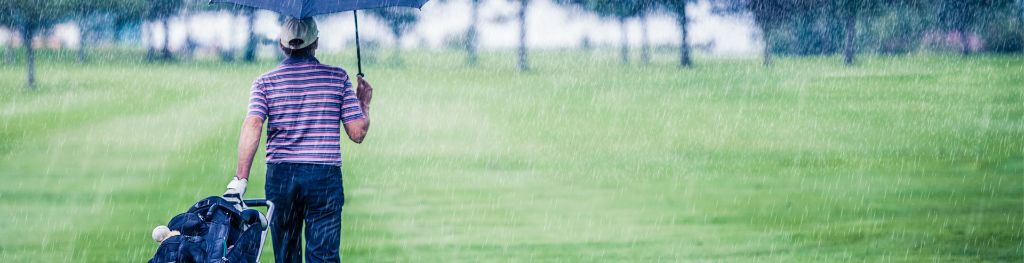 A golfer holding an umbrella walks on a golf course in heavy rain.
