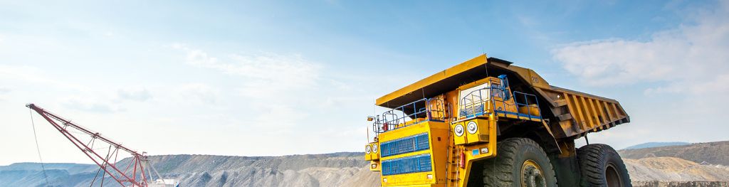 A big yellow mining truck in an opencast mine under a blue sky, and a crane in the background.