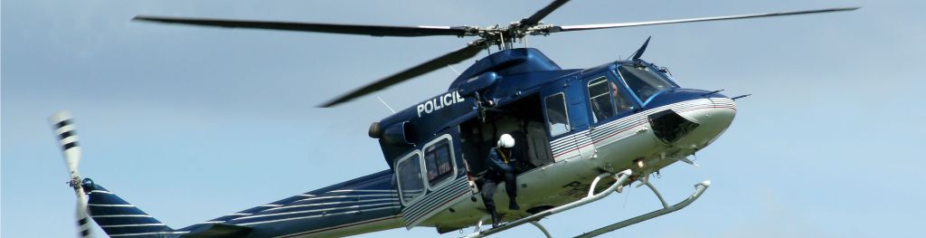 A blue and white police helicopter with an officer suspended in a harness by the door.
