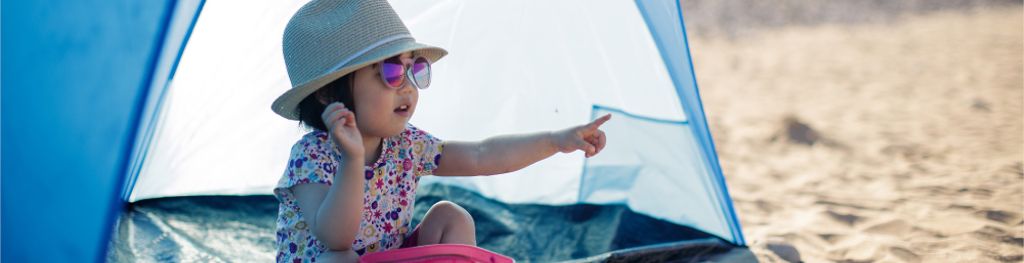 A little girl wearing a hat and sunglasses sits under a protective tent on the beach