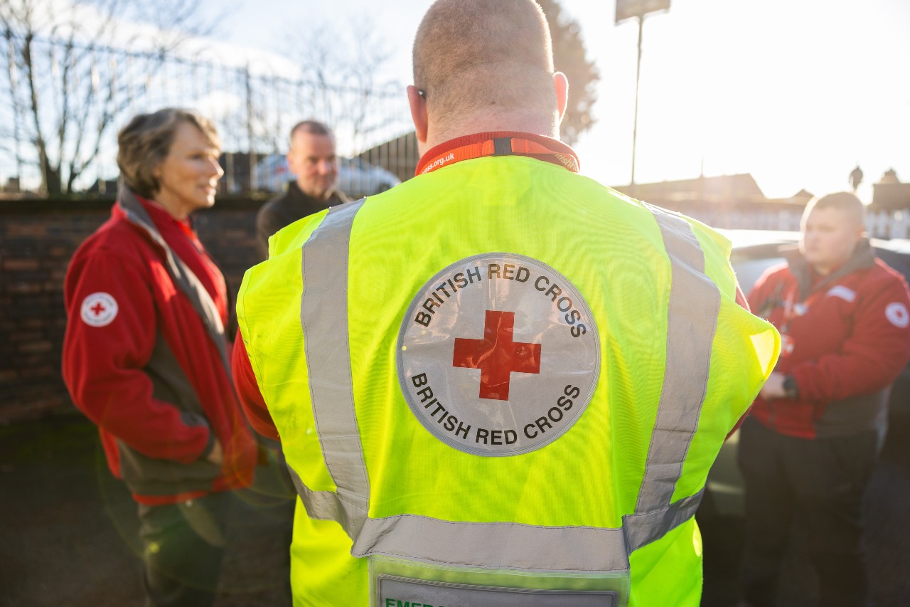 Responders at the site of a flood event in the UK. Image shows the back of a responder wearing a high vis vest with the British Red Cross logo on the back talking to their team.