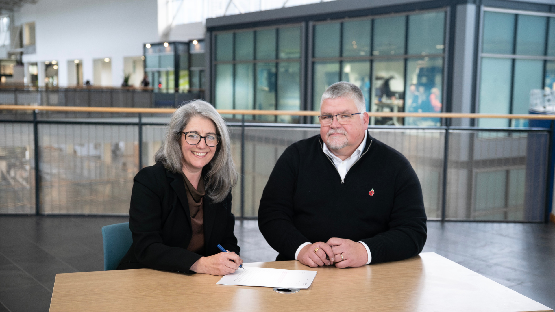 A photo showing the Met Office's Steve Calder and NESO's Deborah Pettersen signing a document at a table. They smile as they hold pens.