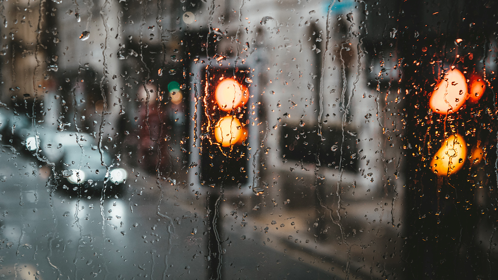 Raindrops on a window with blurred traffic lights and a road in the background.