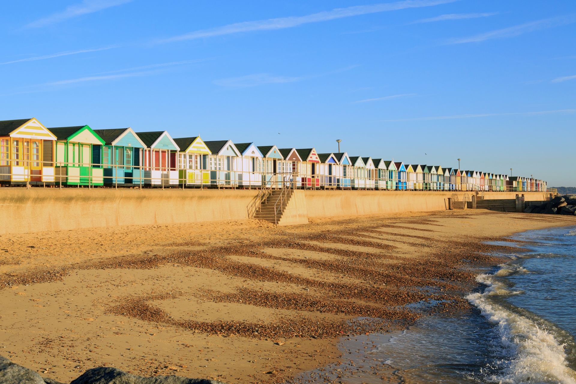 Colourful beach huts on a sandy beach with a wave breaking onto the sand in the right side of the image.